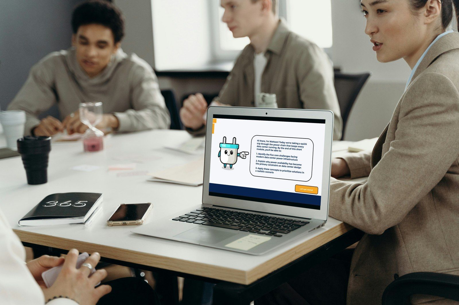 A team at a meeting table with a laptop showing Wattson introducing the Powering the Data Center microlearning
